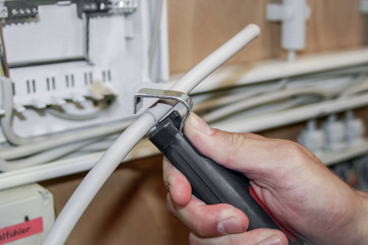 Eine Person schneidet ein weißes Kabel mit einem Kabelschneider in einem Schaltschrank. Im Hintergrund sind Elektroteile sichtbar.