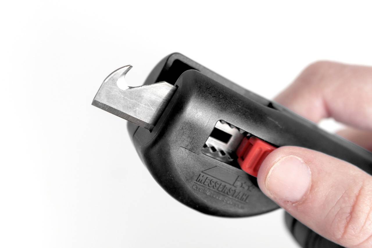 Person hält einen schwarzen Sicherheitskartonöffner mit einem herausragenden Metallmesser, bereit zum Schneiden.