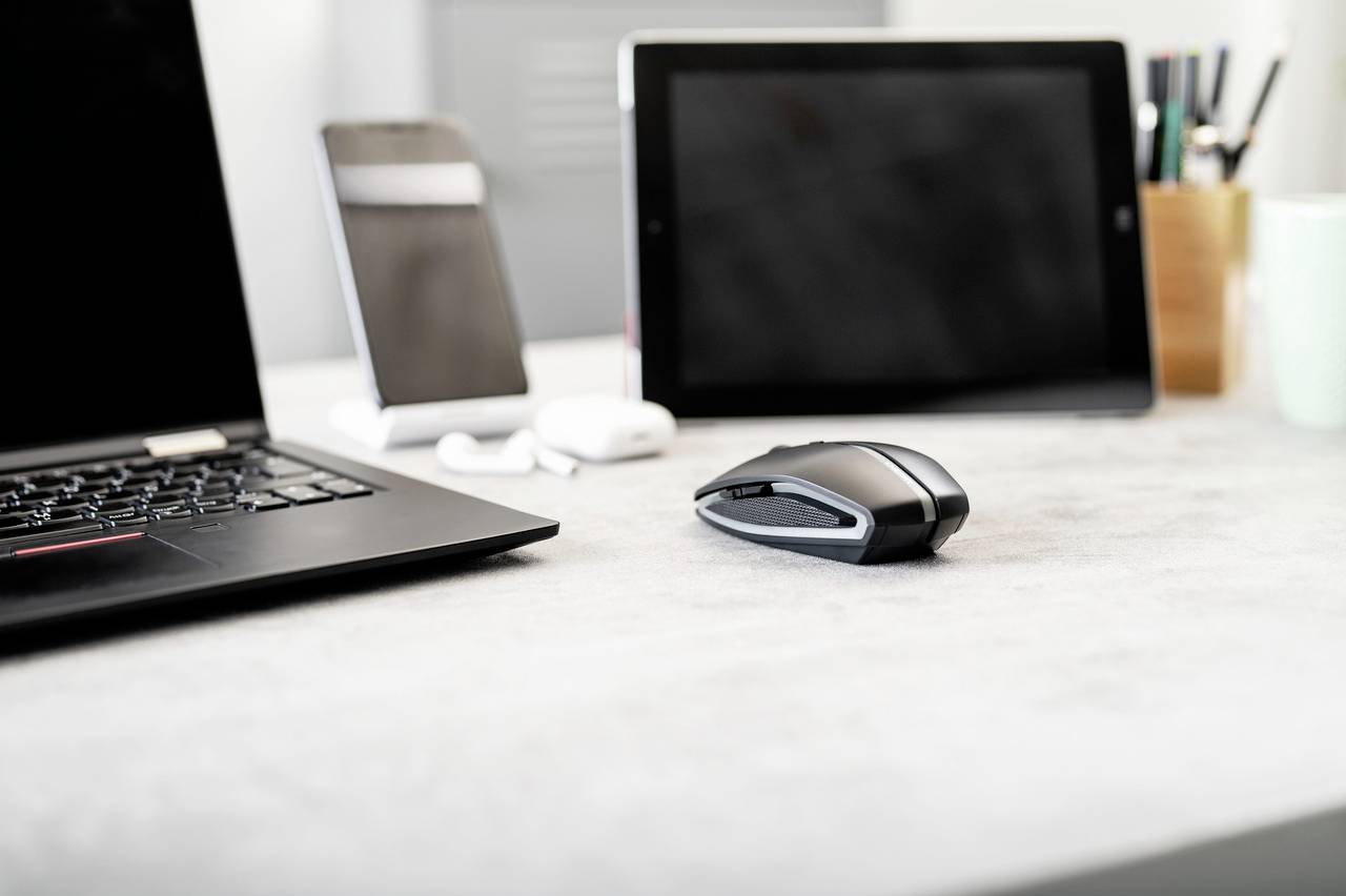 Laptop, Maus, Tablet und Smartphone auf einem Schreibtisch. Geräte sind ausgeschaltet. Büro-Umgebung mit Schreibutensilien im Hintergrund.