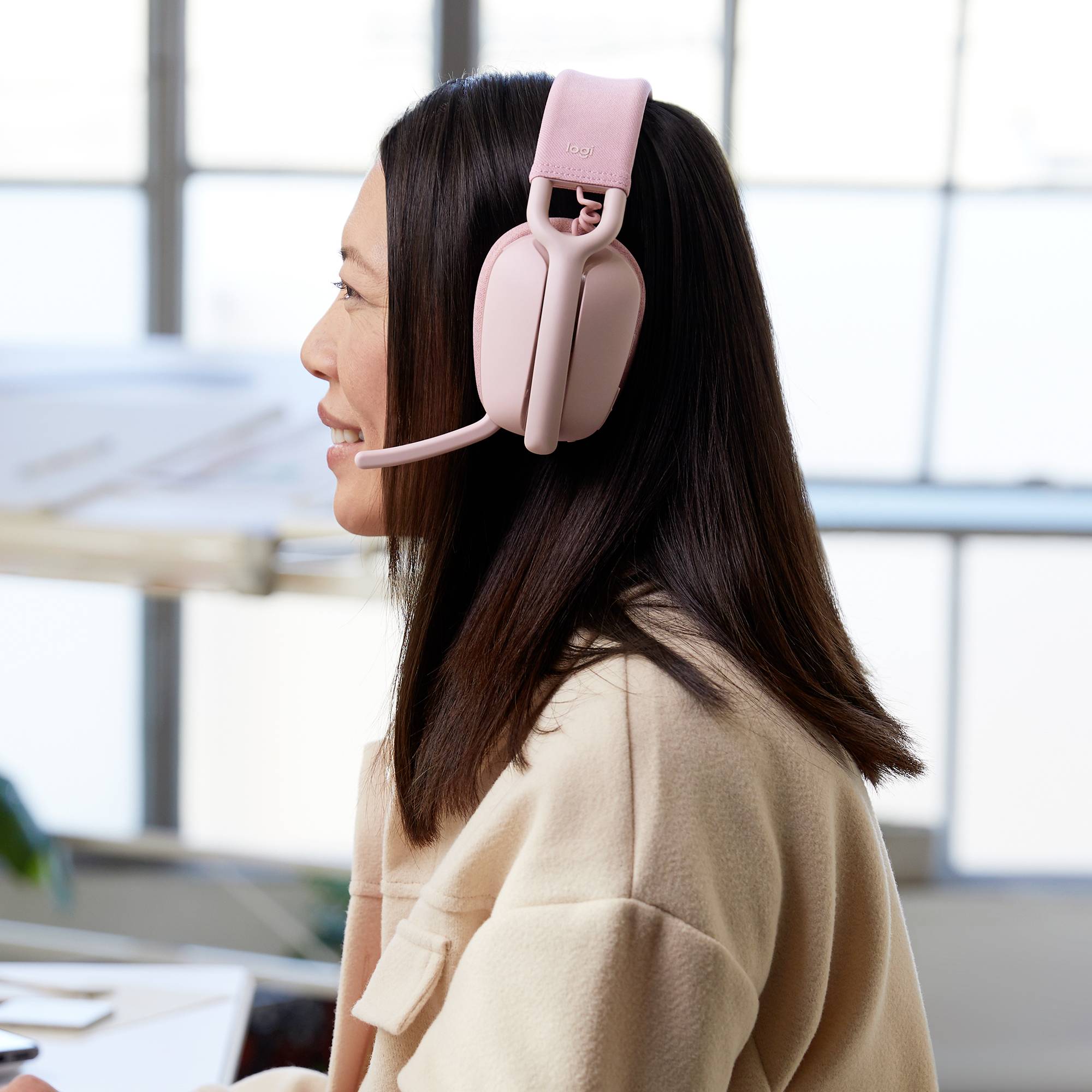 Eine Frau mit langen Haaren trägt rosa Kopfhörer mit Mikrofon und sitzt an einem Schreibtisch. Im Hintergrund ist ein Fenster zu sehen.