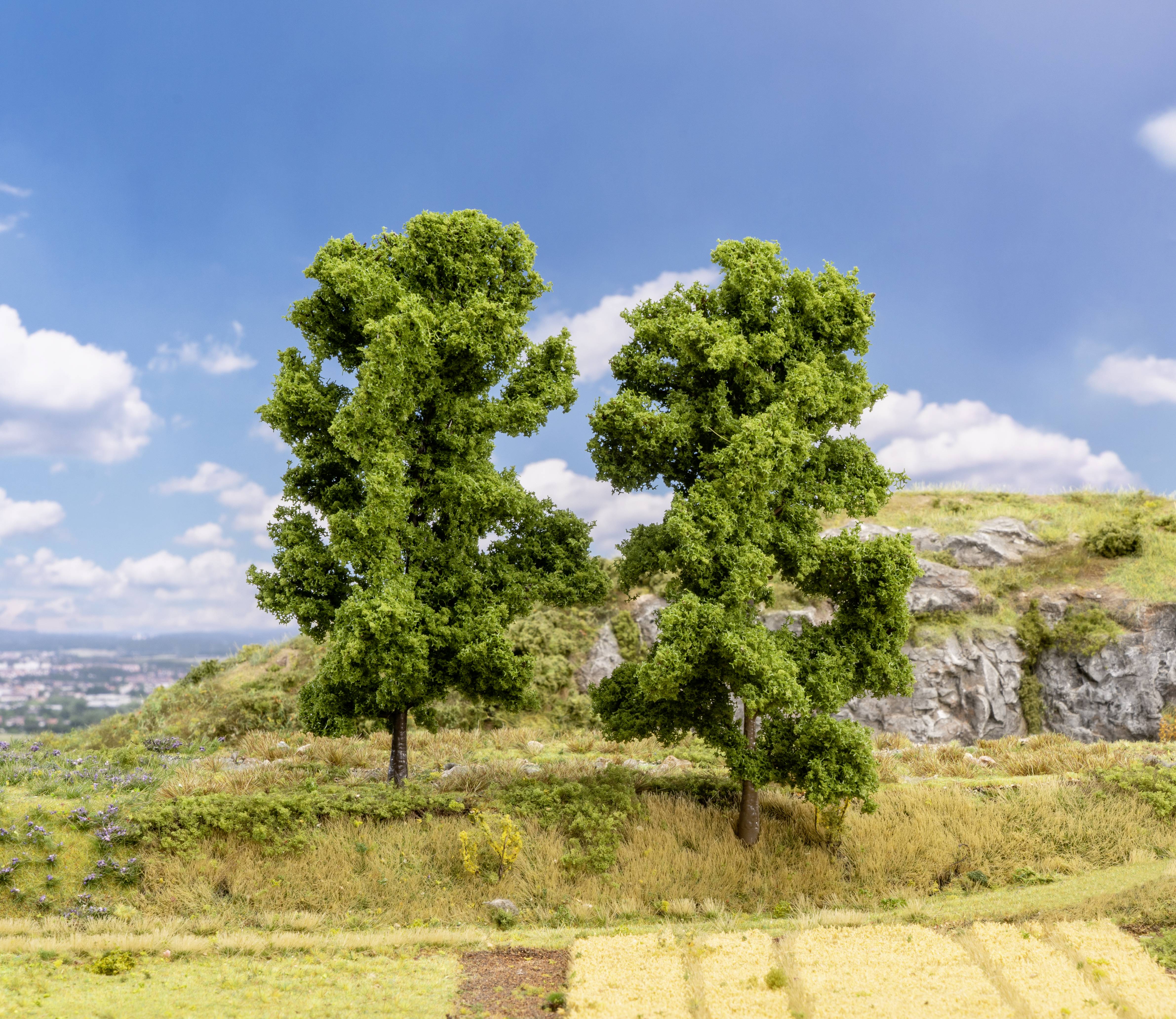 Zwei grüne Bäume auf einem Hügel, umgeben von Gras und Felsen, mit blauem Himmel und Wolken im Hintergrund.