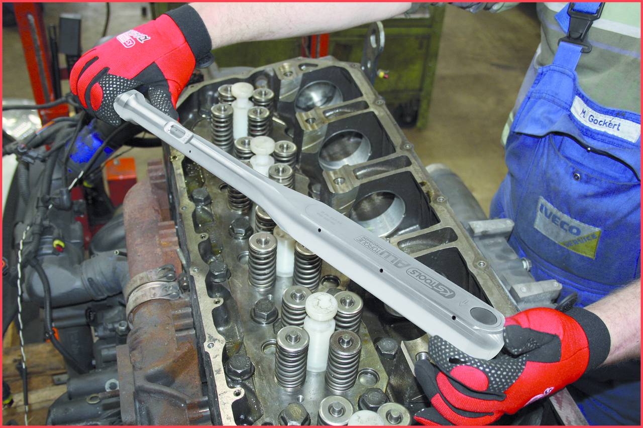 Eine Person in Arbeitskleidung hält ein langes Werkzeug über einem offenen Motorblock mit sichtbaren Ventilen.