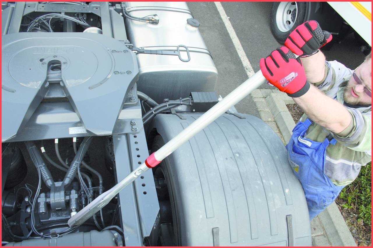 Eine Person überprüft die Mechanik eines LKW-Anhängers mit einem Werkzeug. Sie trägt rote Handschuhe und steht neben den Reifen.