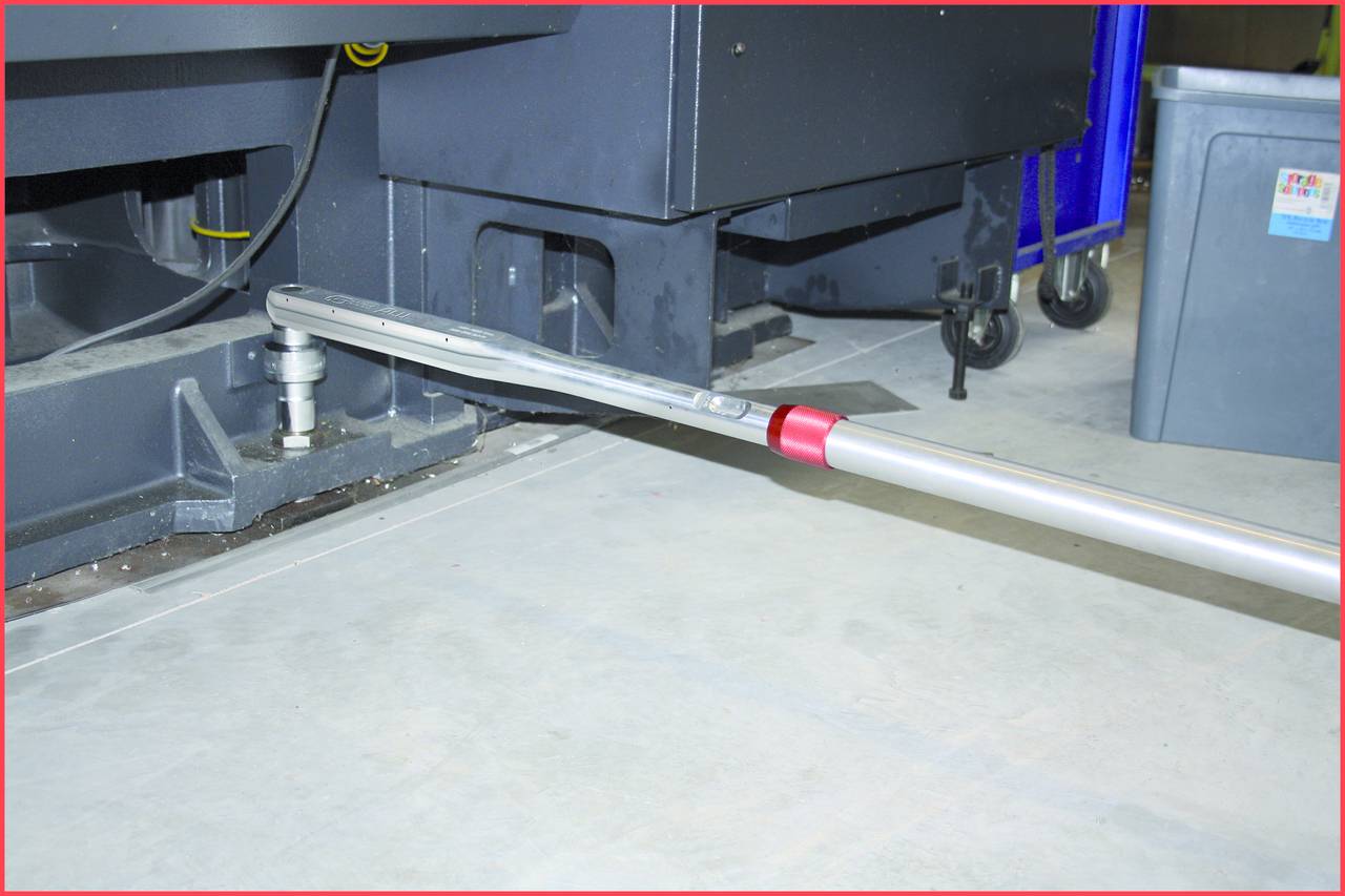 Ein langer Metallstab ist unter einer großen Maschine am Boden platziert. Rechts steht ein grauer Behälter im Hintergrund.