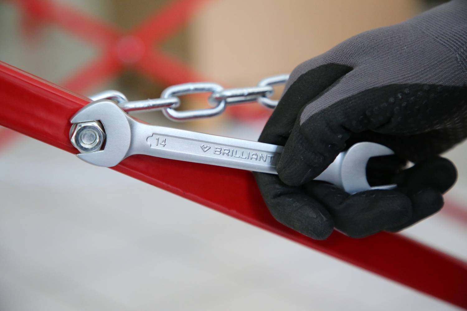 Ein Handschuh hält einen Schraubenschlüssel, der eine Schraube an einem roten Metallgestell anzieht. Eine Kette ist am Rahmen befestigt.