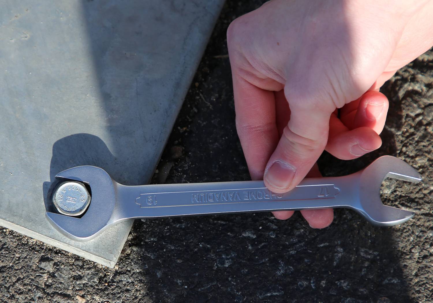 Eine Hand hält einen Schraubenschlüssel, der eine Schraube festhält, auf einer asphaltierten Oberfläche neben einer Metallplatte.