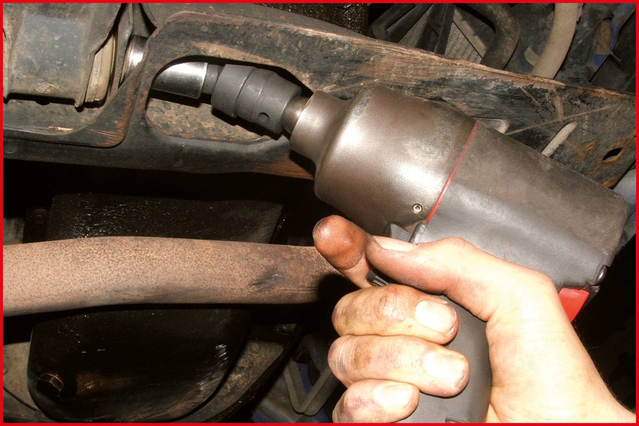 Eine Hand hält eine elektrische Ratsche und zieht eine Schraube an einem Auto fest. Die Umgebung ist schmutzig und industriell.