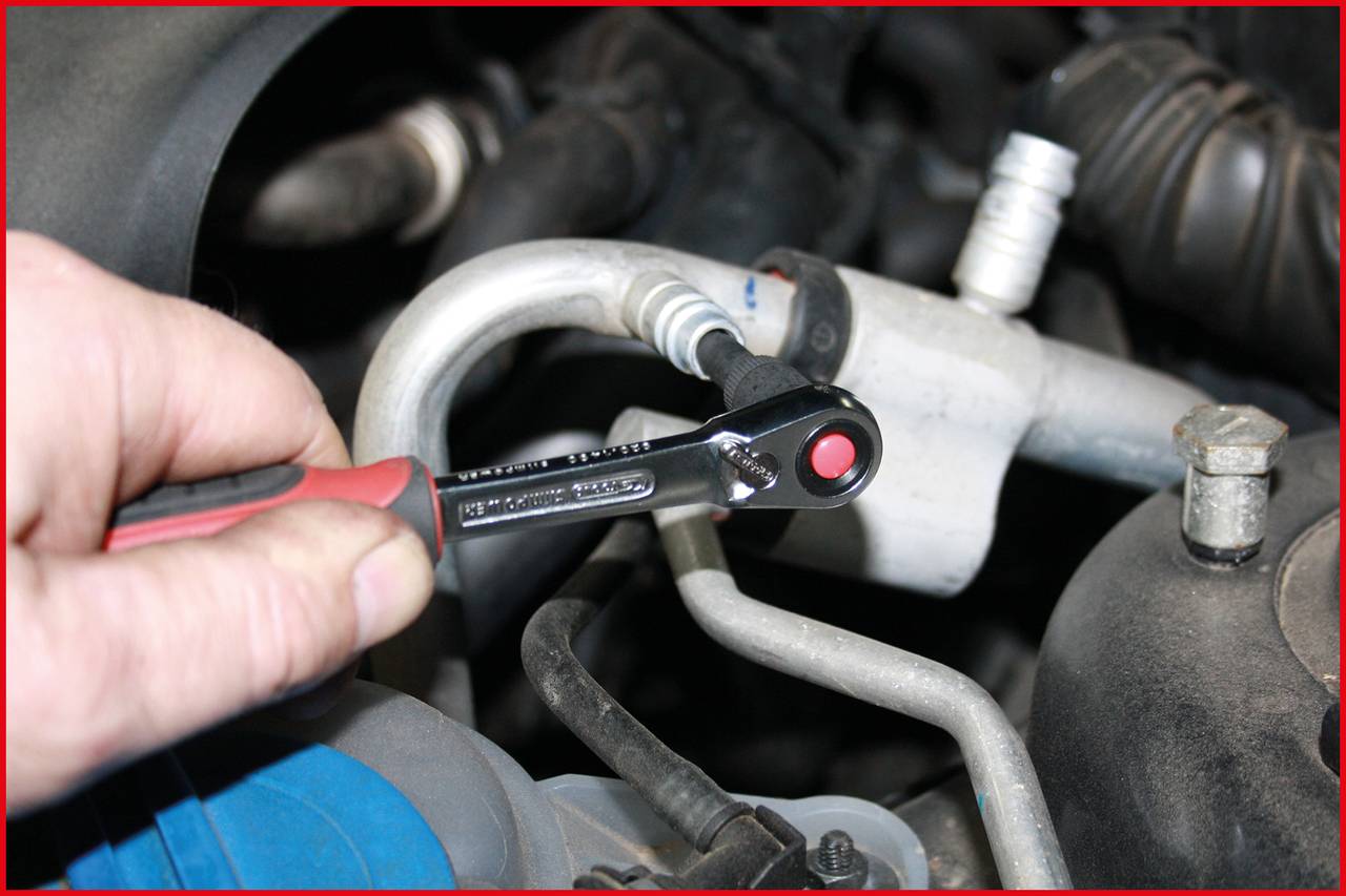 Eine Hand hält einen Ratschenschlüssel und zieht eine Schraube an einem Autoteil an, vermutlich im Motorraum.