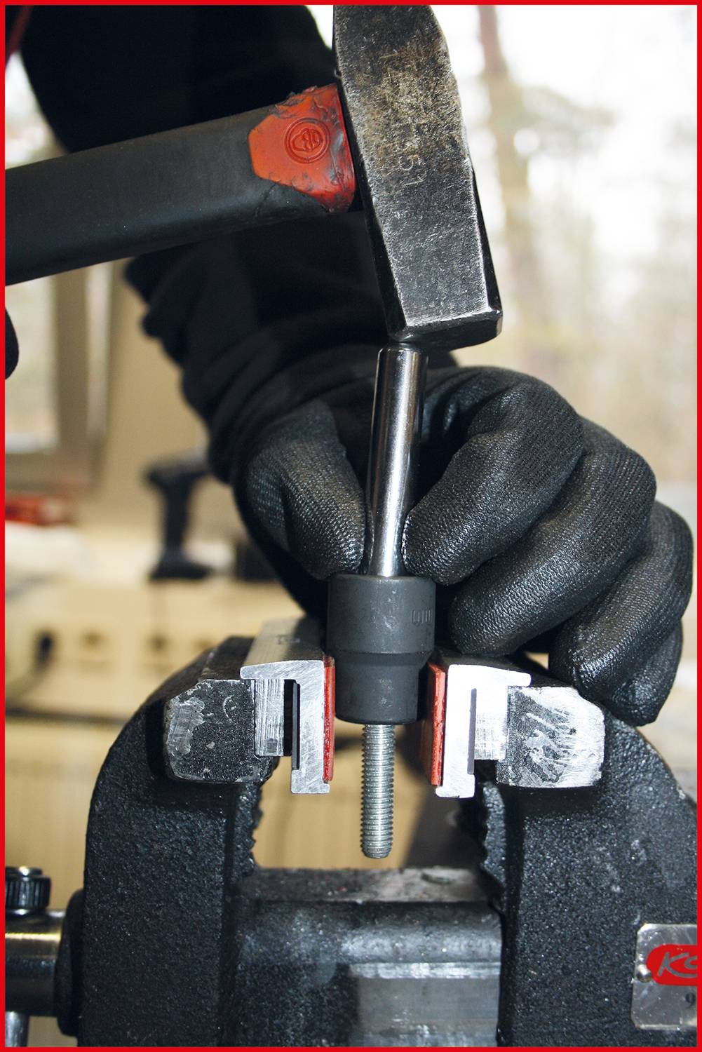 Eine Hand hält einen Metallstab in einem Schraubstock, während ein Hammer ihn bearbeitet. Werkstattumgebung im Hintergrund.