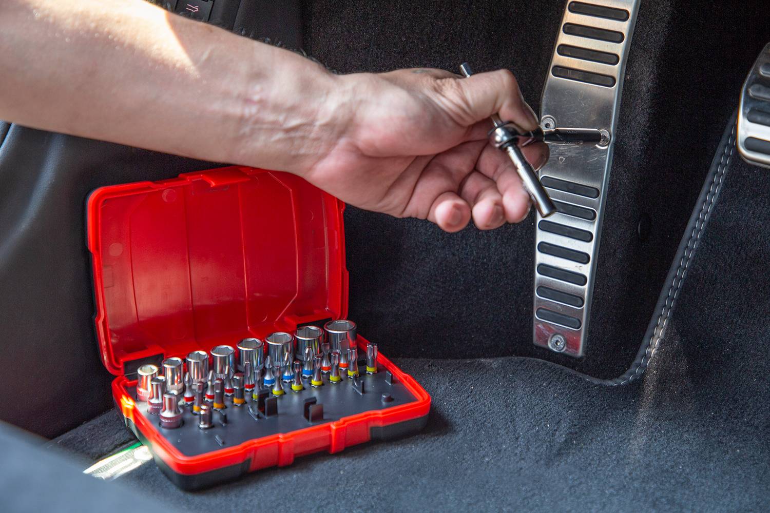 Eine Hand verwendet ein Schraubenwerkzeug, um eine Schraube in einem Auto zu befestigen. Ein rotes Werkzeugset liegt daneben auf dem Boden.