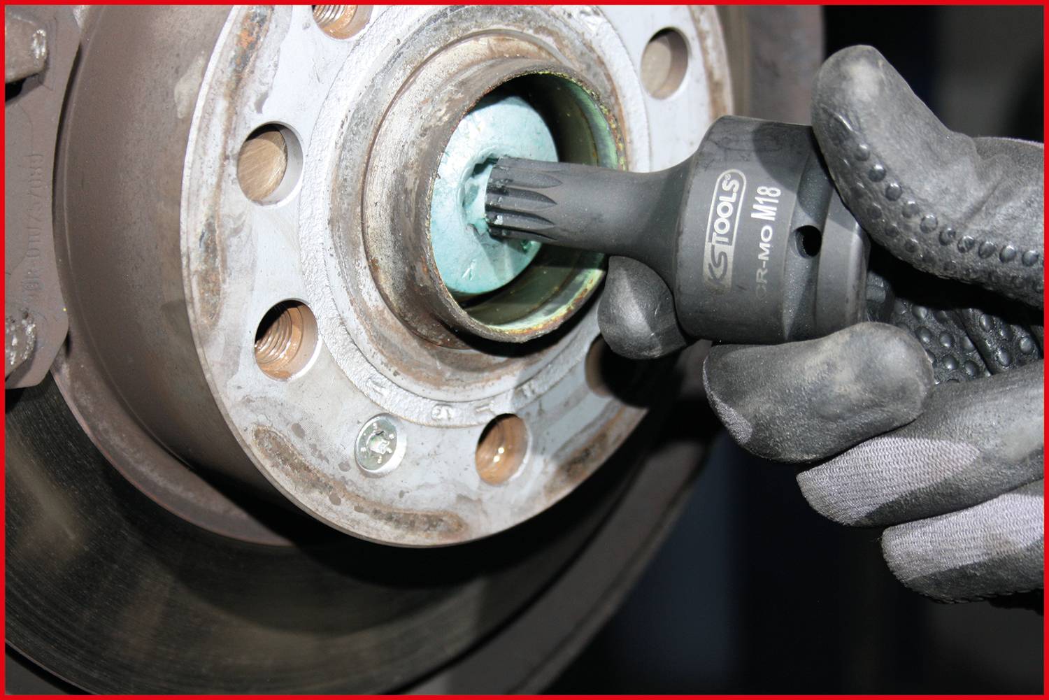 Eine Hand mit Arbeitshandschuh hält einen Werkzeugaufsatz und arbeitet an einem Autoteil, wahrscheinlich an einer Bremsscheibe.