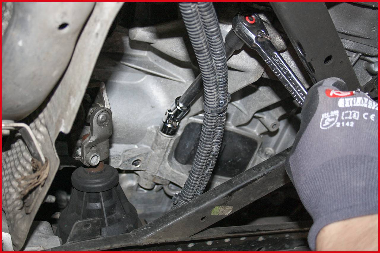 Eine Hand mit Handschuh hält einen Ratschen-Schlüssel und zieht eine Schraube an einem Fahrzeugmotor an. Kabel und Motorteile sichtbar.