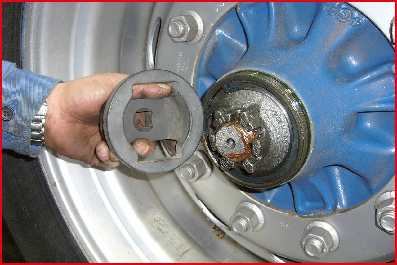 Eine Hand hält eine schwarze Abdeckung neben einem blauen Radnabenbauteil mit sichtbaren Schraubenmuttern und Bolzen.