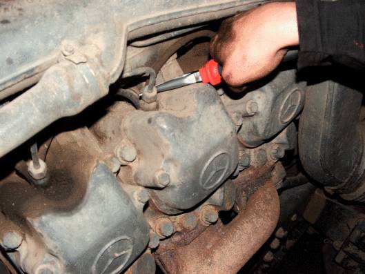 Eine Hand mit Schraubendreher repariert den Motor eines Fahrzeugs. Der Motor ist schmutzig und zeigt Gebrauchsspuren.