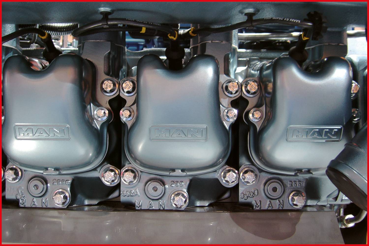 Ein Motorblock von MAN mit drei nebeneinander liegenden silbernen Zylinderabdeckungen, sichtbar sind Verschraubungen und Verkabelung.