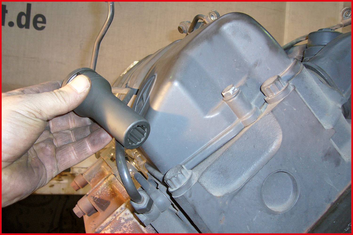 Eine Hand hält ein spezielles Werkzeug, das auf einen Bolzen einer grau lackierten Maschine oder Motorteils aufgesetzt wird.
