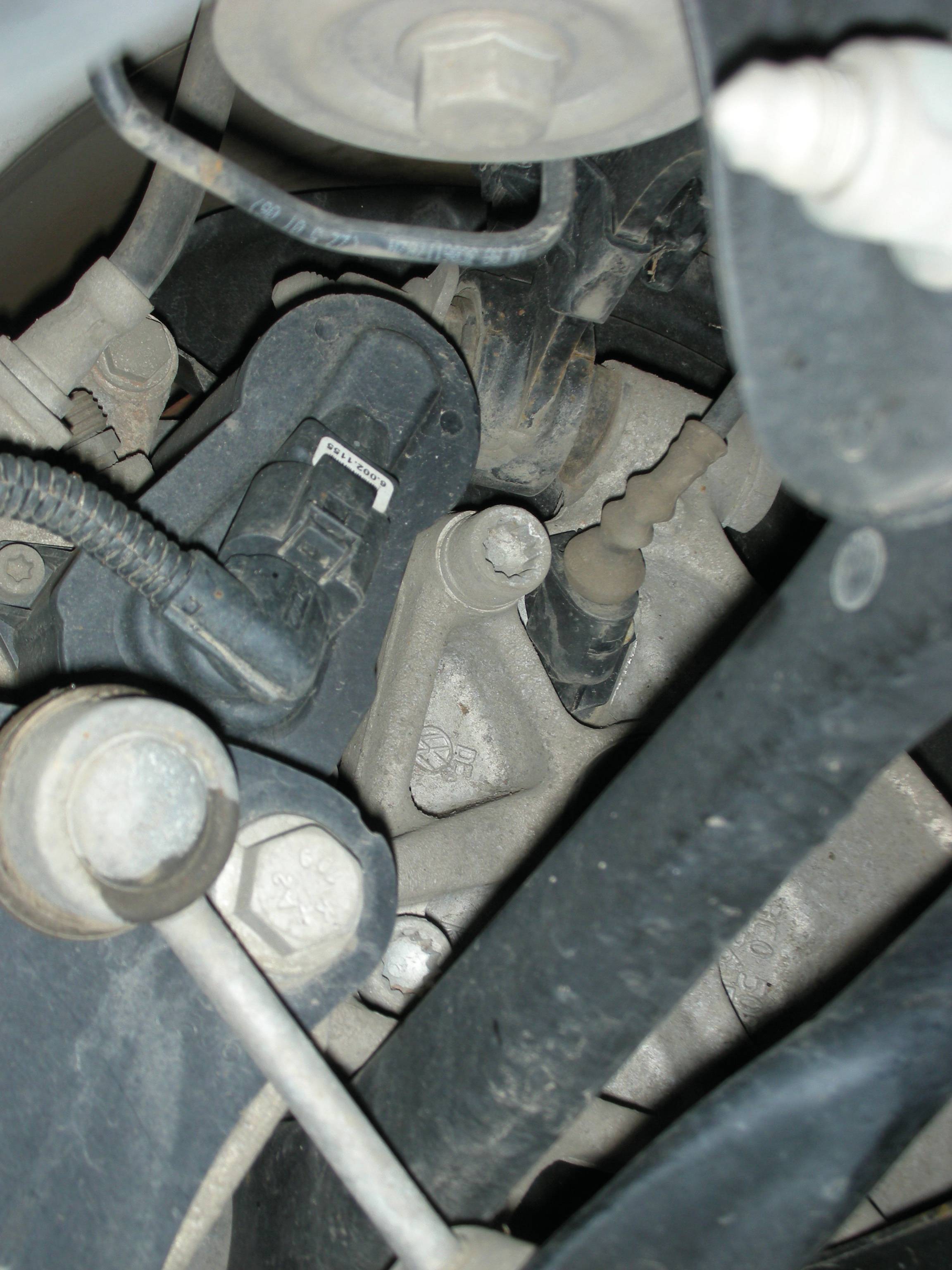 Nahaufnahme von Autoteilen im Motorraum. Verschiedene Kabel, Schrauben und Metallkomponenten sind sichtbar.