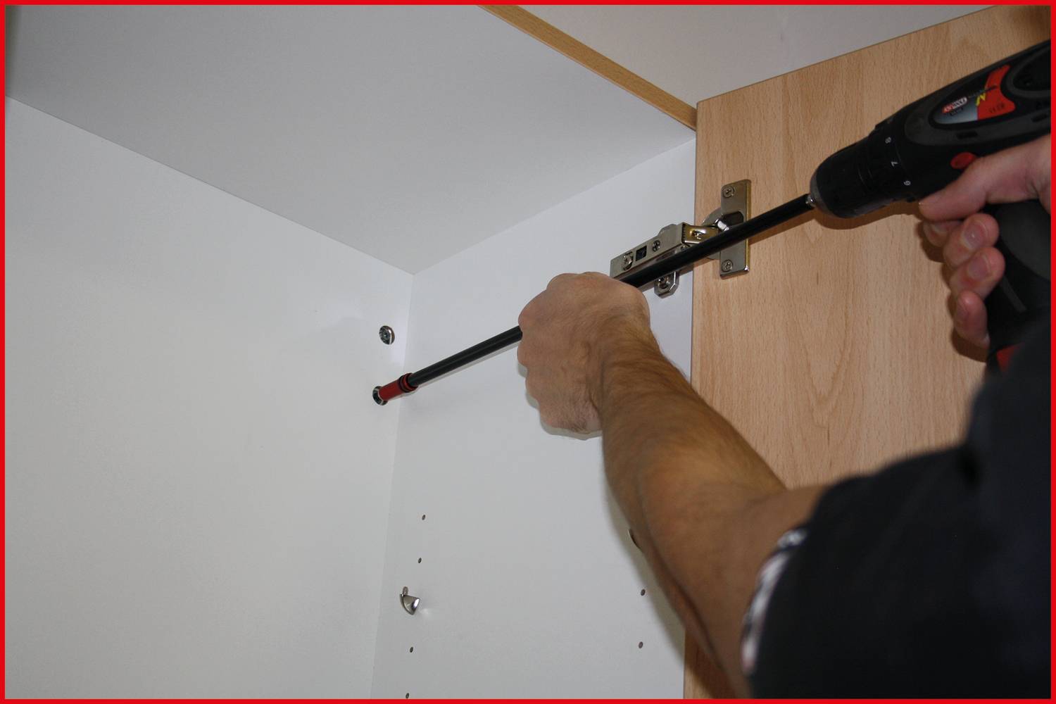 Eine Person befestigt ein Scharnier an einem Holzschrank mit einem schwarzen Akkuschrauber in einem weißen Innenraum.