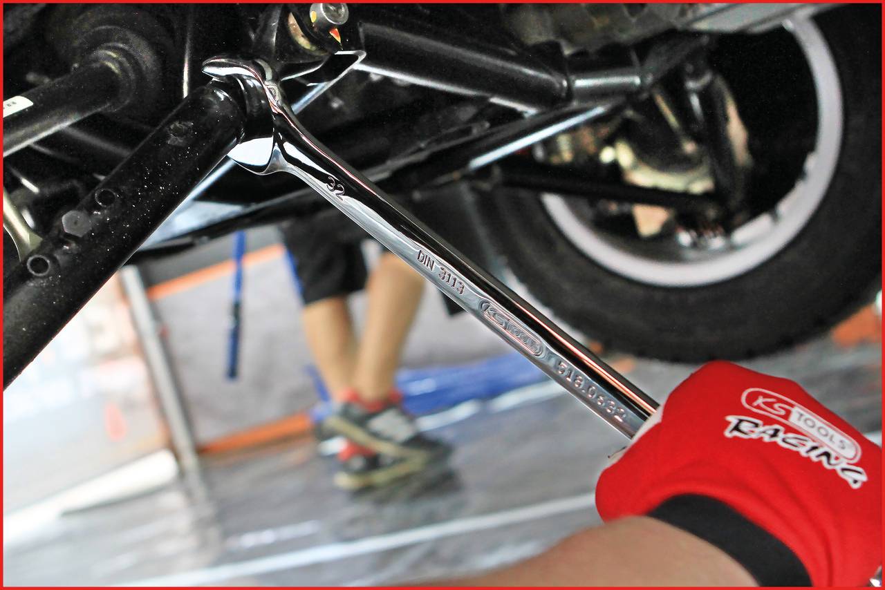 Ein Mechaniker repariert ein Fahrzeug mit einem Schraubenschlüssel. Der Fokus liegt auf dem Werkzeug, während die Hände in roten Handschuhen sichtbar sind.