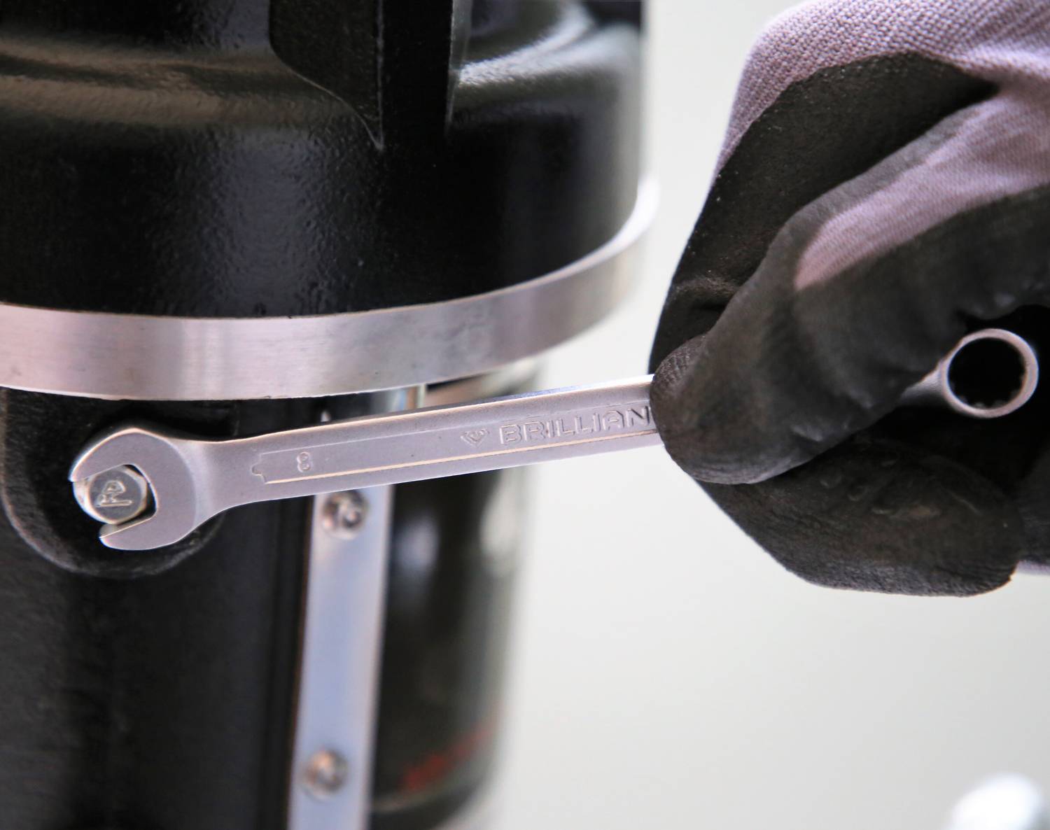 Ein Handschuh hält einen Schraubenschlüssel, der an einer Schraube eines Metallgeräts angesetzt ist. Gezeigt wird eine Wartungsarbeit.