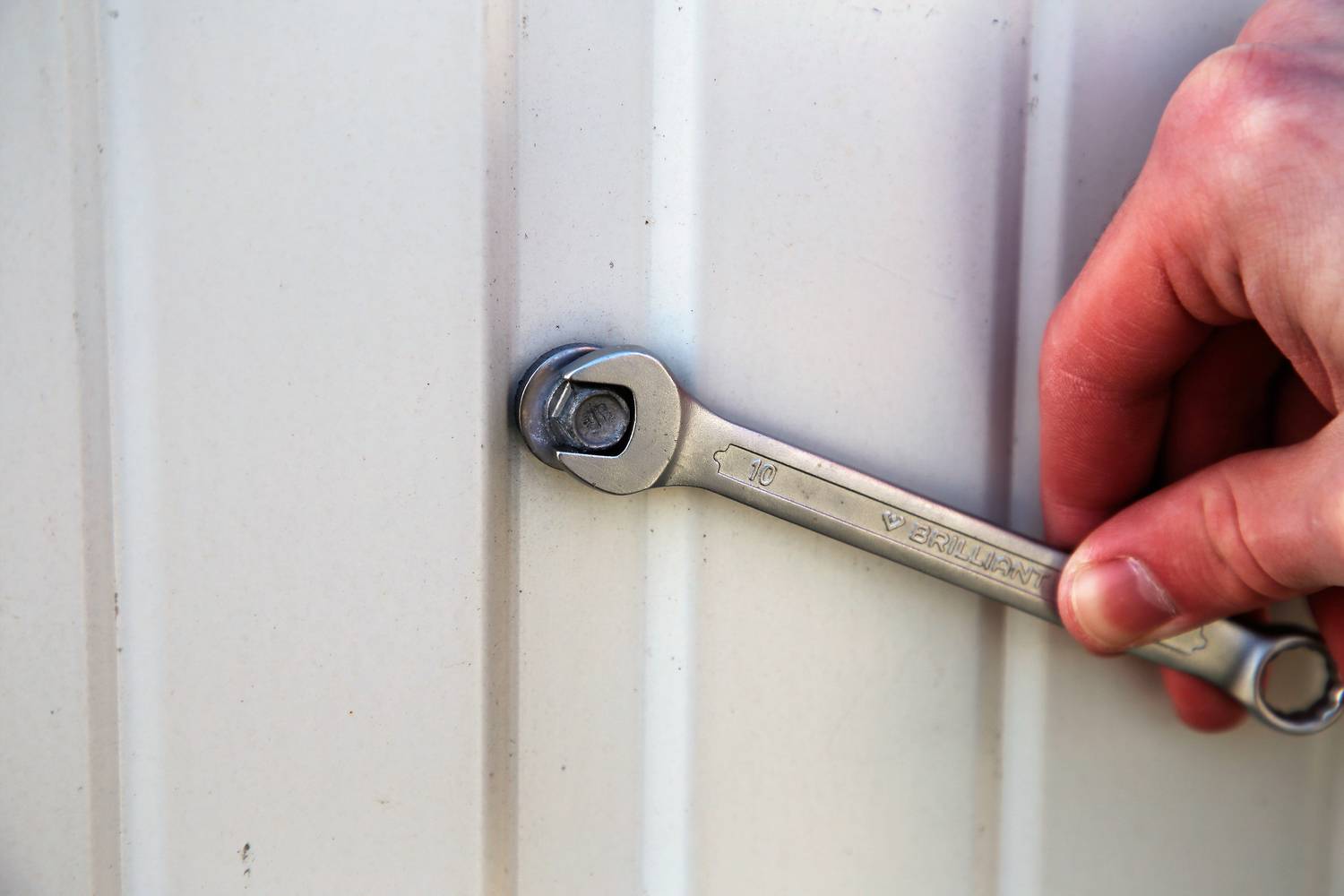 Eine Hand zieht mit einem Schraubenschlüssel eine Schraube an einem weißen, gerippten Metallblech fest.