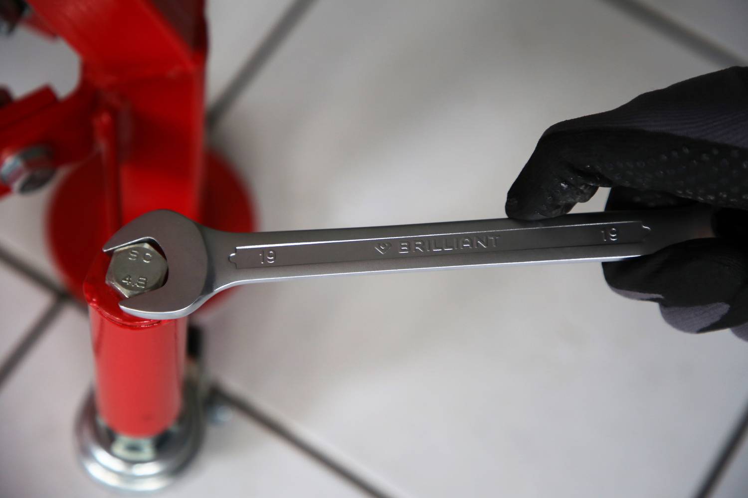 Eine Hand mit Handschuh hält einen offenen Ringschlüssel neben einem roten Metallobjekt auf gefliesten Boden.