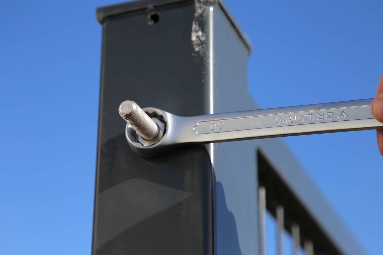 Eine Hand hält einen Schraubenschlüssel, um eine Metallmutter an einem vertikalen Zaunpfosten festzuziehen, blauer Himmel im Hintergrund.