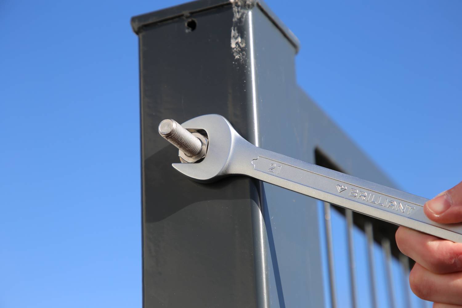 Eine Hand zieht mit einem Schraubenschlüssel eine Schraube an einem Metallpfosten an, während der Himmel im Hintergrund blau ist.