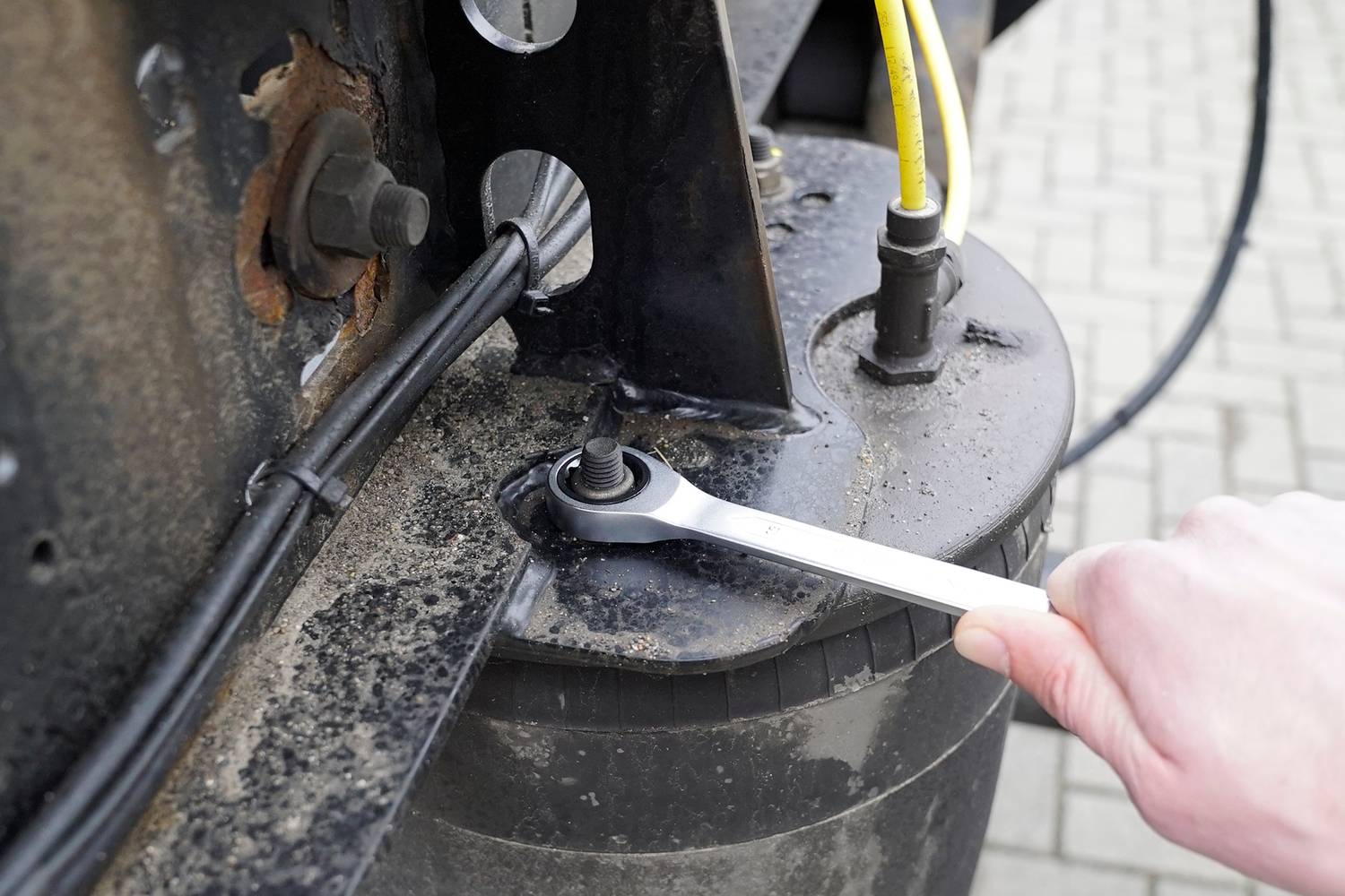 Hand zieht mit Schraubenschlüssel eine Schraube an einem metallischen Teil fest. Im Hintergrund sind Pflastersteine zu sehen.