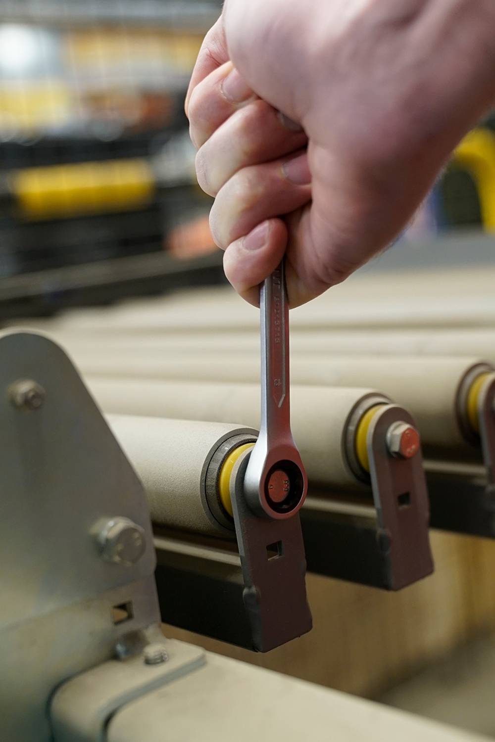 Eine Hand hält einen Schraubenschlüssel, der an einer Maschine arbeitet. Die Maschine hat mehrere Rollen und befindet sich in einer Werkstattumgebung.