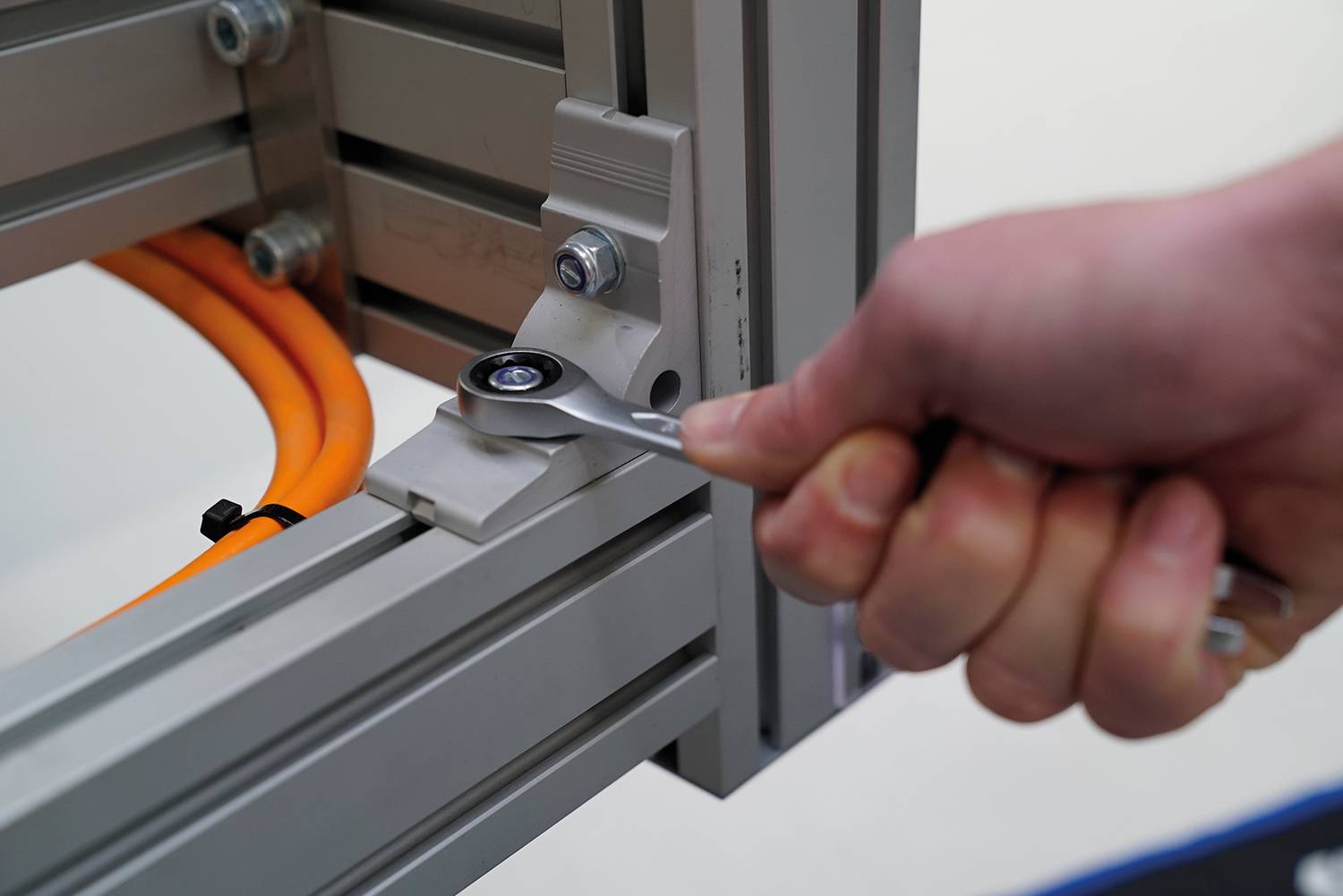 Hand zieht mit einem Schraubenschlüssel eine Schraube an einem metallenen Rahmengestell fest. Im Hintergrund sind orangefarbene Kabel sichtbar.