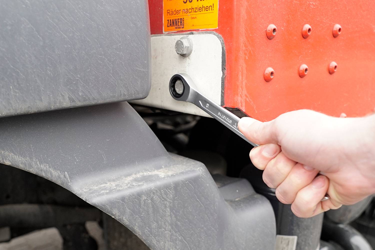 Eine Hand hält einen Schraubenschlüssel und schraubt an einer Schraube eines orange-grauen Landfahrzeugs.
