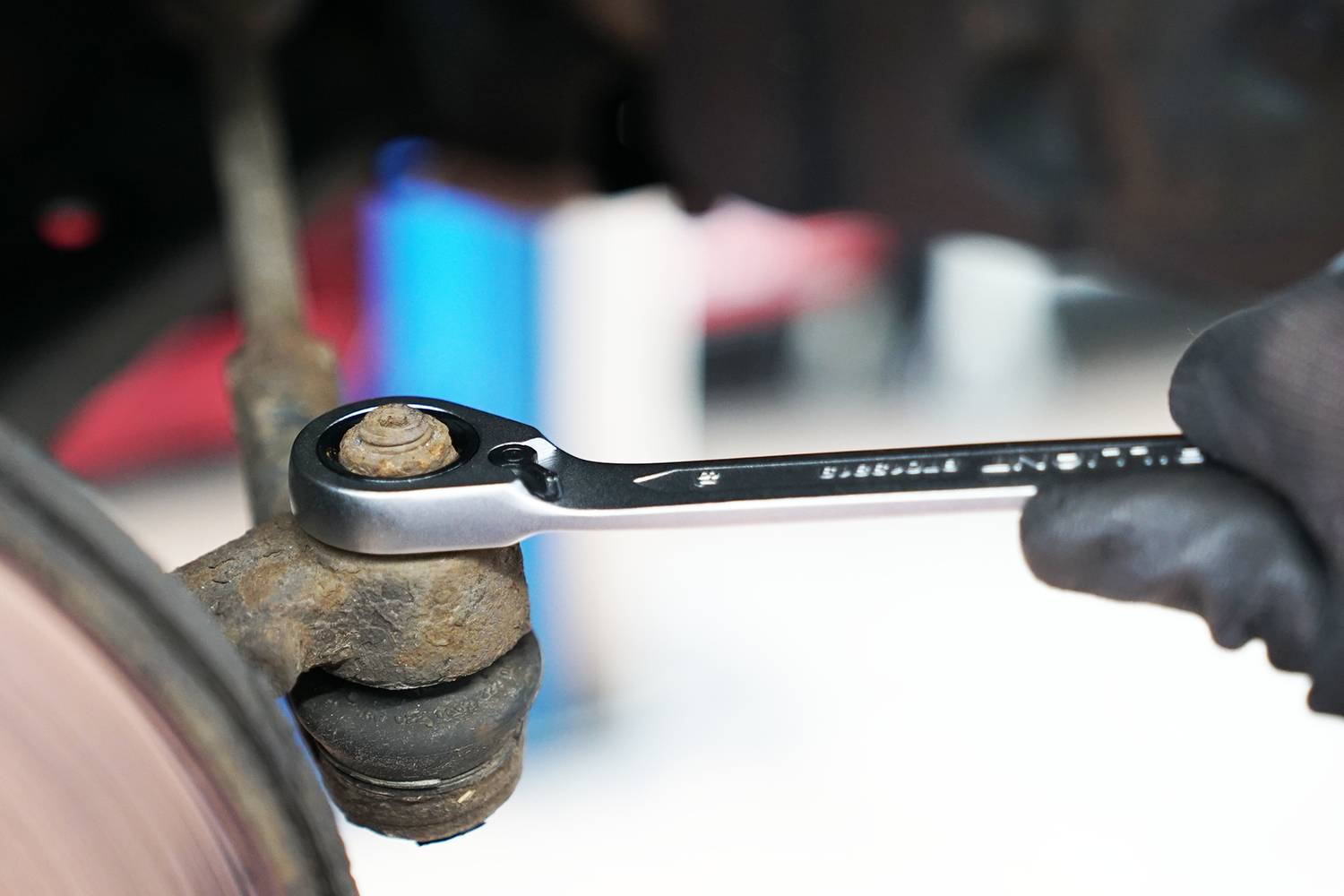 Eine Hand hält einen Schraubenschlüssel und zieht eine Schraube an einem Autoteilen fest. Im Hintergrund sind unscharfe Werkstattdetails zu sehen.