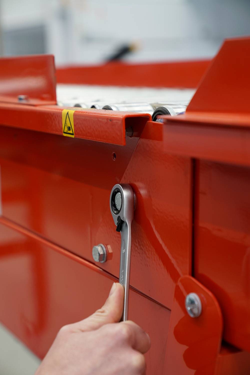 Eine Person benutzt einen silbernen Schraubenschlüssel, um eine rote Industriemaschine anzupassen. Fokus auf Hand und Werkzeug.