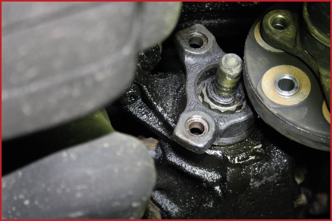 Nahaufnahme einer dunklen, verölten Autokomponente mit zwei Befestigungslöchern und einer zentralen Schraube; Teil eines Fahrzeugmotors.