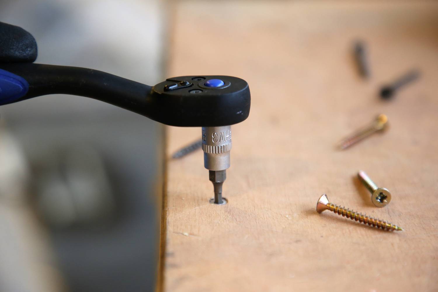 Ein Akkuschrauber zieht eine Schraube in eine Holzplatte ein. Mehrere Schrauben liegen daneben auf der Oberfläche verstreut.