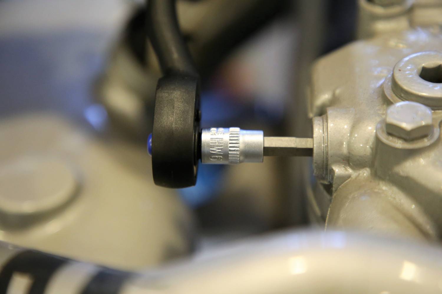 Nahaufnahme eines Metallventils an einem Maschinen- oder Fahrzeugteil mit einem schwarzen Schlauch und Schraube im Hintergrund.