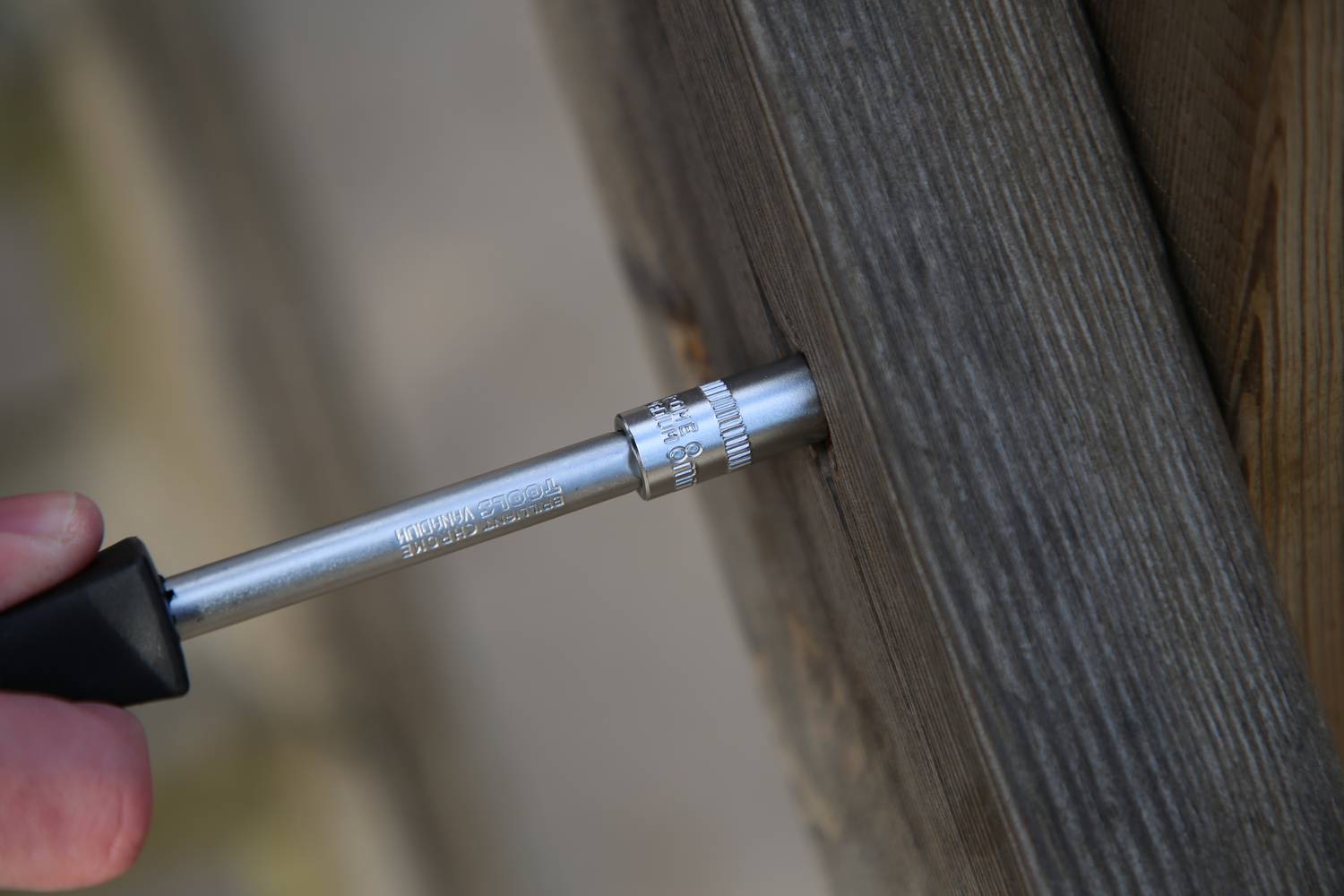 Eine Person misst die Tiefe eines Bohrlochs in einem Holzbalken mit einem Tiefenmesser. Der Balken erscheint dunkelbraun und texturiert.