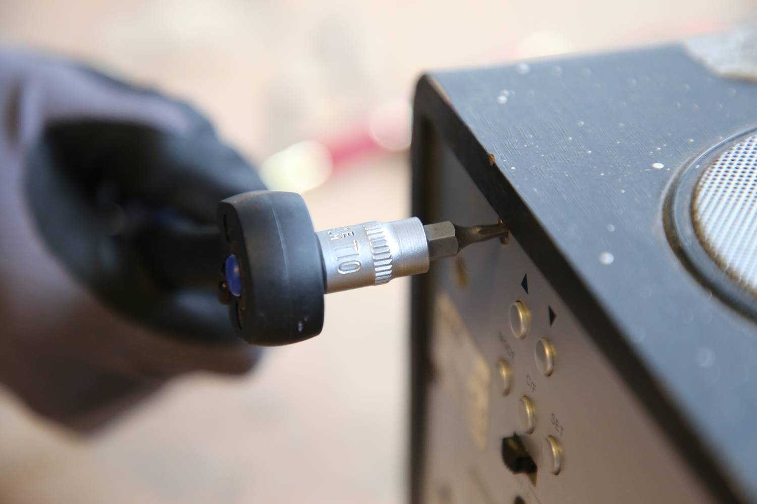 Eine Person mit Handschuhen zieht mit einem Werkzeug eine Schraube an einem elektronischen Gerät fest.