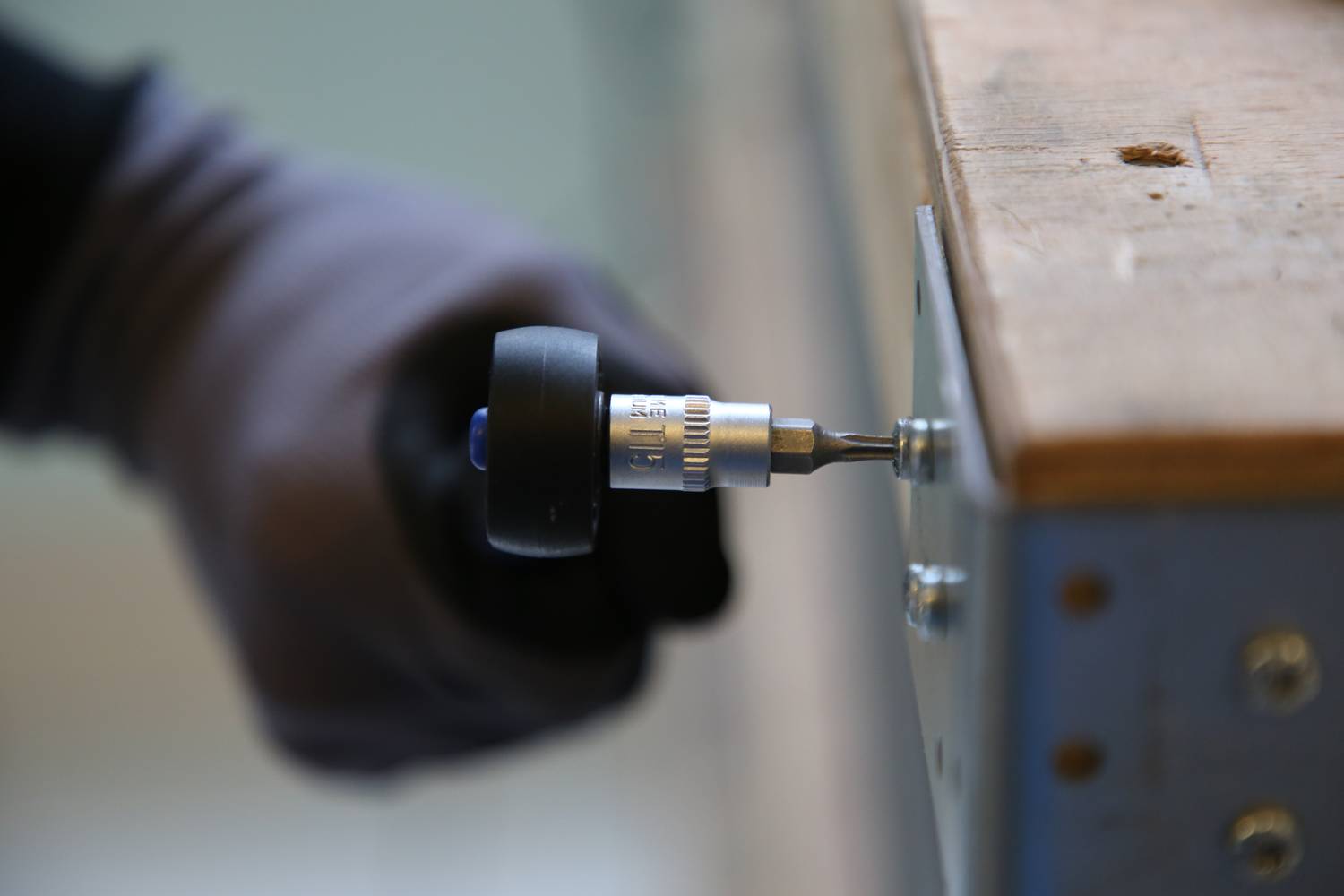 Eine Person zieht mit einem Akkuschrauber eine Schraube an einem Holztisch fest. Der Fokus liegt auf der Schraube und dem Werkzeug.