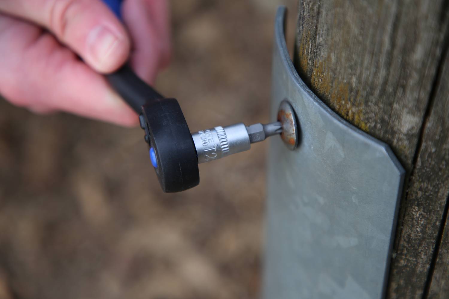 Eine Hand zieht mit einem Schraubenschlüssel eine Schraube an einem Metallblech fest, das an einem Holzpfosten befestigt ist.