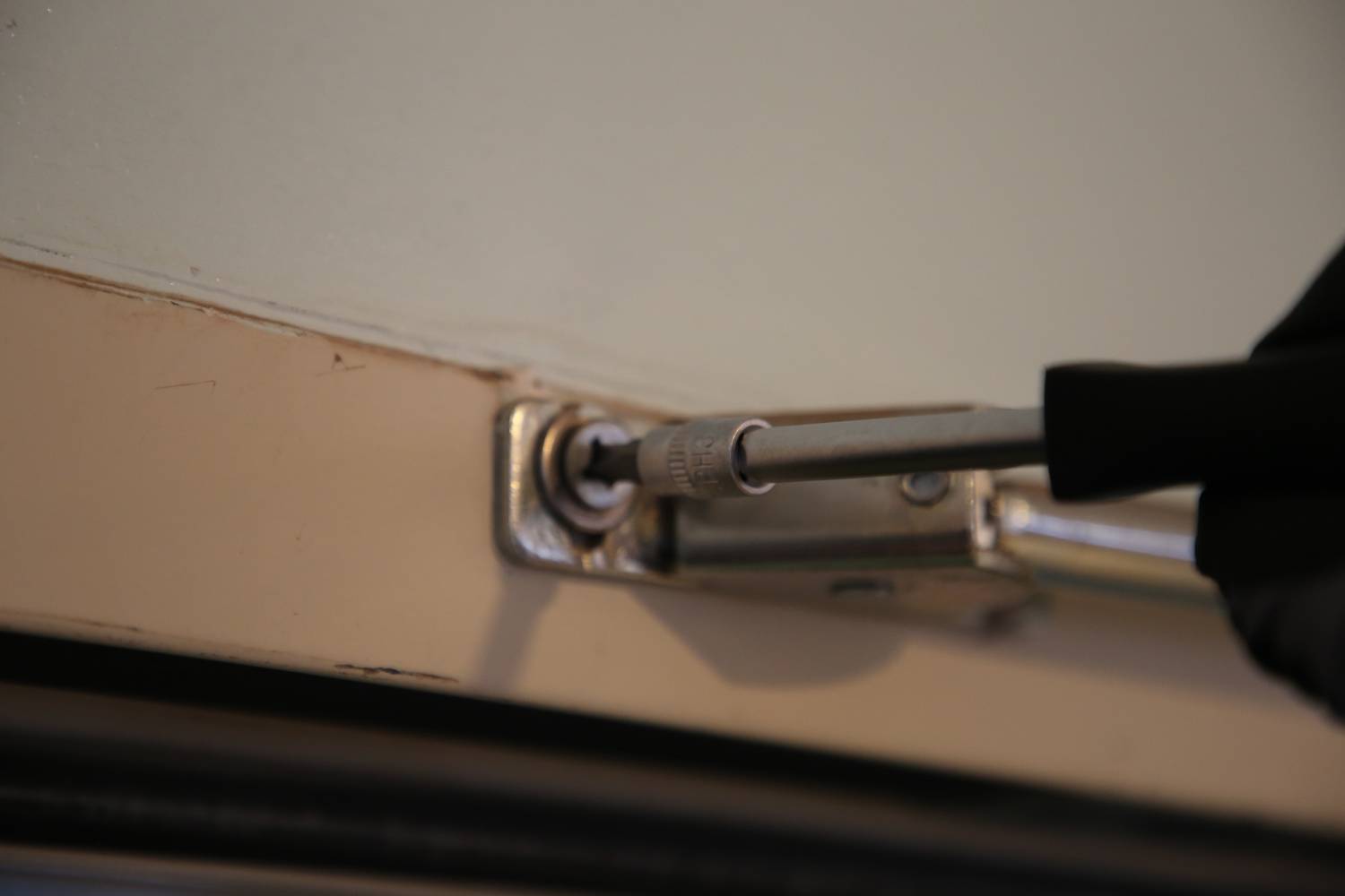 Eine Hand mit Handschuh dreht eine Schraube in einem Türscharnier fest, um die Tür zu reparieren oder zu justieren.