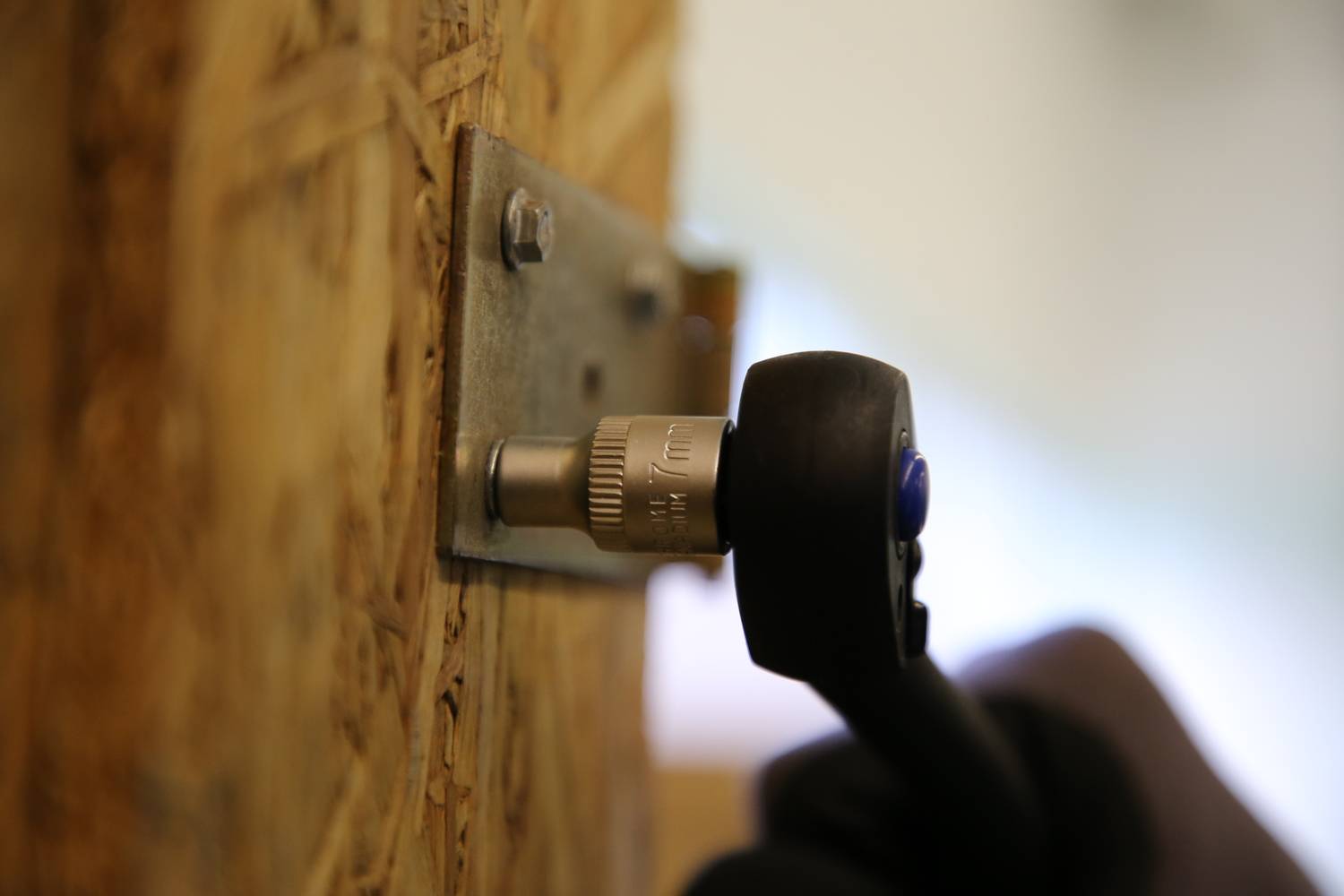 Hand dreht mit Ratsche eine Schraube in ein Holzbrett. Schraubenschlüssel auf Metallplatte fixiert. Nahe Aufnahme.