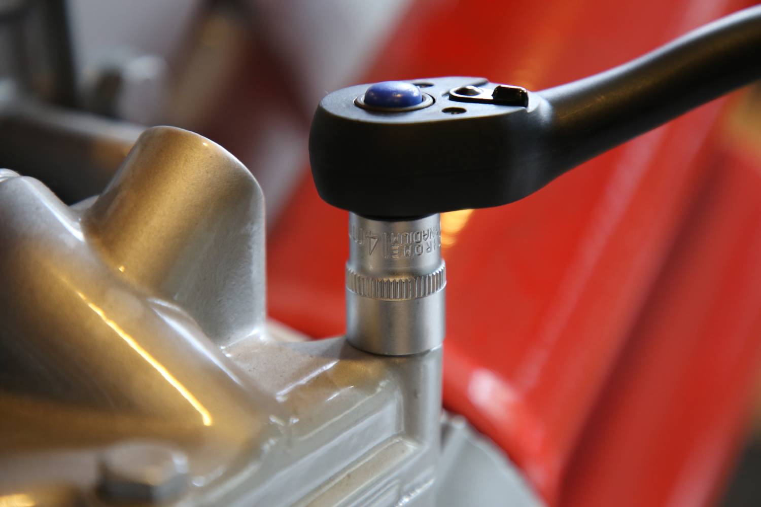 Eine Ratsche wird auf eine Schraube an einer silbernen Maschinenkomponente angewendet, im Hintergrund ist ein verschwommener roter Teil sichtbar.