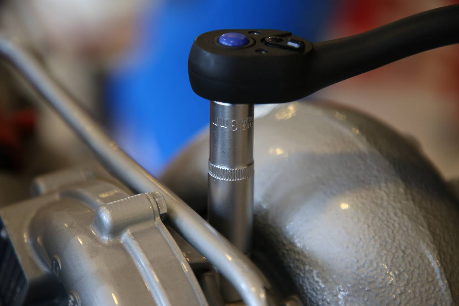 Ein Drehmomentschlüssel zieht eine Schraube an einem metallischen Motorteil fest. Im Hintergrund unscharfe blaue und rote Flächen.