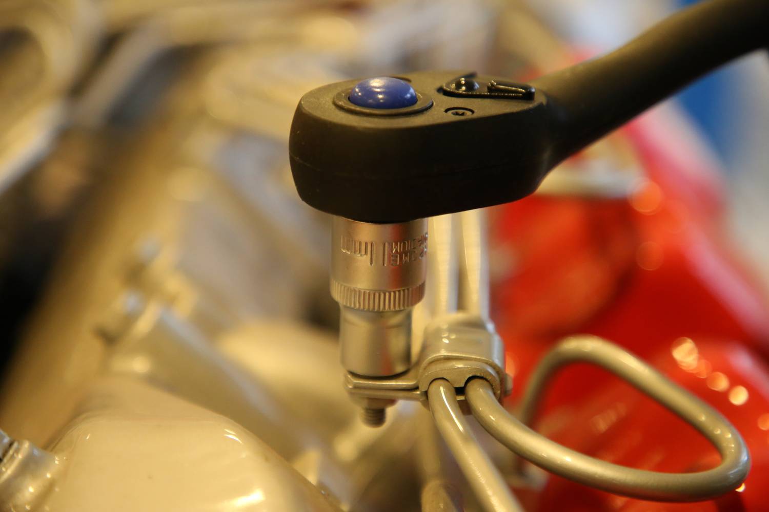 Eine Ratsche zieht eine Schraube auf einem Motor an. Das Werkzeug hat einen blauen Knopf und ist auf ein metallisches Teil gerichtet.