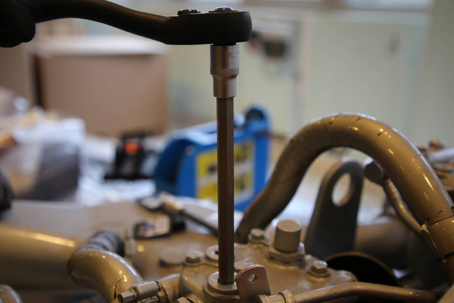 Eine Person benutzt ein Werkzeug, um eine Schraube an einem Automotor anzuziehen. Im Hintergrund sind unscharfe Werkstattobjekte.