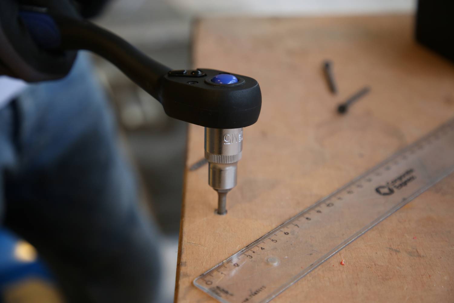 Eine Person bohrt ein Loch in ein Holzbrett mit einem elektrischen Bohrer. Daneben liegt ein durchsichtiger Lineal mit Markierungen.
