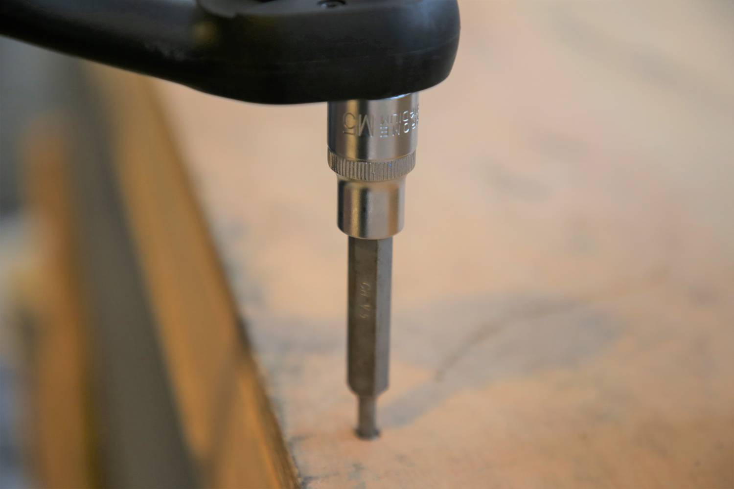 Ein Bohrer bohrt ein Loch in ein Holzbrett. Im Hintergrund sind unscharfe Details einer Werkbank zu erkennen.