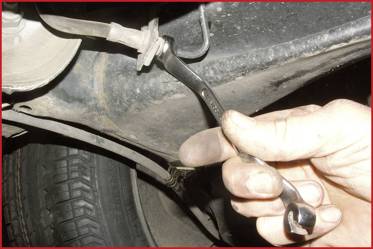 Eine Hand hält einen Schraubenschlüssel, um an einem Auto zu arbeiten. Ein Reifen und ein Teil der Fahrzeugunterseite sind sichtbar.